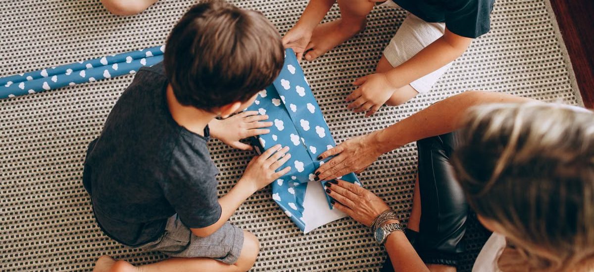 Kids wrapping a gift with blue paper featuring white clouds on a woven rug.