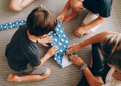 Kids wrapping a gift with blue paper featuring white clouds on a woven rug.